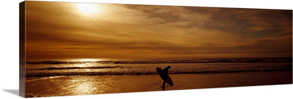 This horizontal panoramic photograph shows a lone figure emerging from the surf as the sun sinks below the horizon.