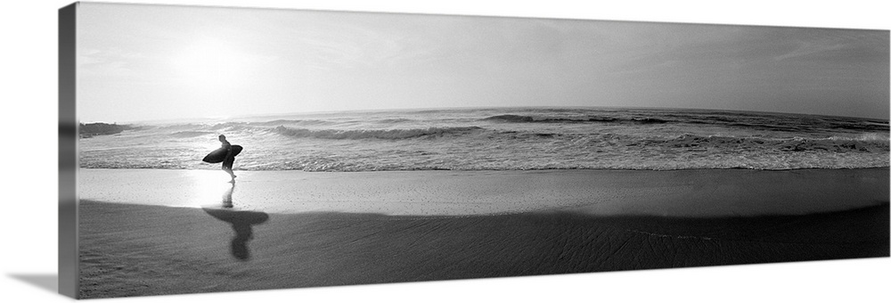 Panoramic photograph of a surfer walking along a sandy beach in San Diego, California.  The tide of the ocean gently crash...
