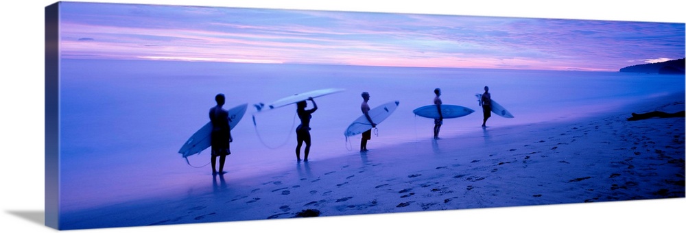 Panoramic photograph shows a group of five surfboarders walking along a sandy shoreline under dim light in Central America.