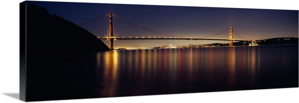 Wide angle photograph of the Golden Gate Bridge in the distance, lit at night and reflecting over the waters of the San Fr...