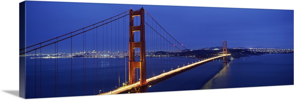Panoramic photo of a bridge spanning the bay, its famous red towers and cables standing out against blue twilight sky.