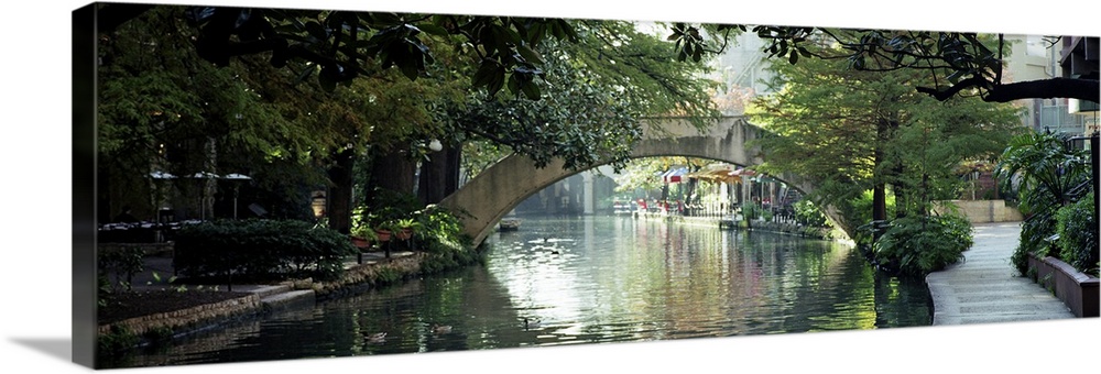 Panoramic image of the canal in San Antonio with sidewalks, trees, and restaurants lining the water and an arched foot bri...