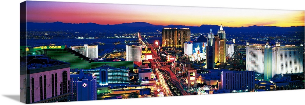 Panoramic photograph of city at sunset with mountains in the distance.  The buildings are brightly lit and a street full o...