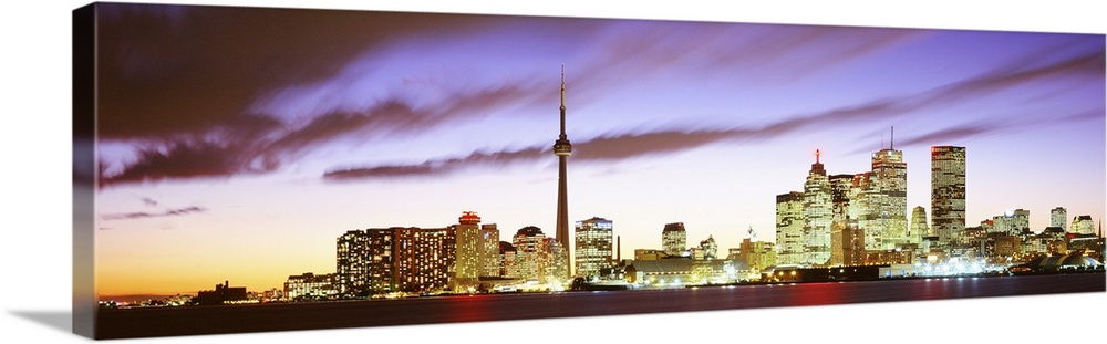 Wide angle photograph of the Toronto skyline, including CN Tower, lit up at dusk, beneath whipping clouds in Ontario, Canada.