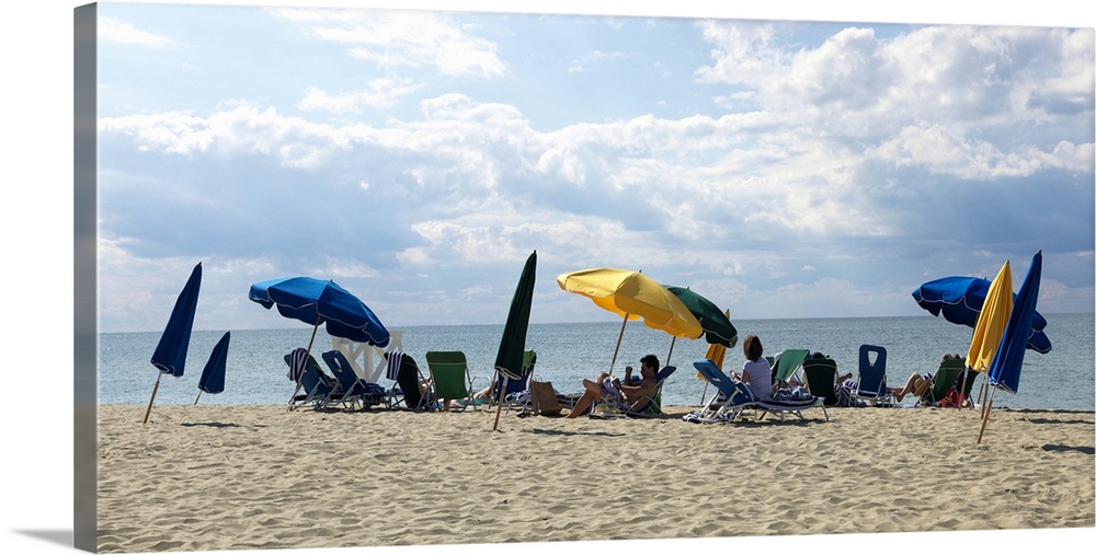 Slanted beach umbrellas shade people that sit on the beach and look out over the ocean.