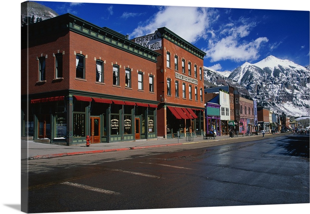 Photograph of street corner lined with buildings and shops with snow covered mountains in the background under a cloudy sky.