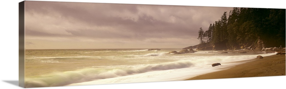 Panoramic picture of waves hitting the shore as the sun peaks through the cloudy sky.