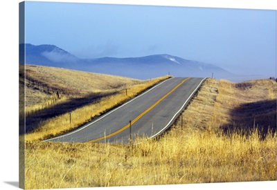 Two lane highway through autumn color grasses, distant mountains in mist, Montana