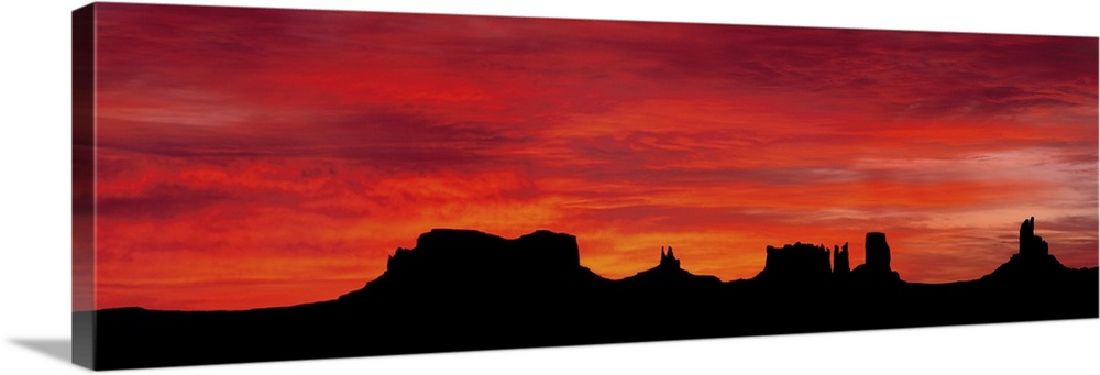Panoramic photograph of silhouettes of huge rock formations in the desert at sunset under a cloudy sky.