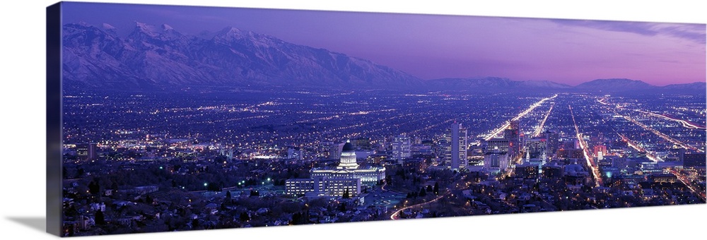 High-angle panoramic photograph of city lit up at dusk with snow covered mountains in the distance under a cloudy sky.