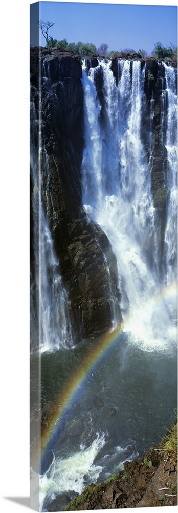 A vertical panoramic piece of an immense waterfall with a rainbow stretching across the bottom of the picture.