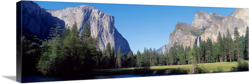 Panoramic photo of large rock cliffs and mountings surrounded by pine trees and water in Yosemite National Park.