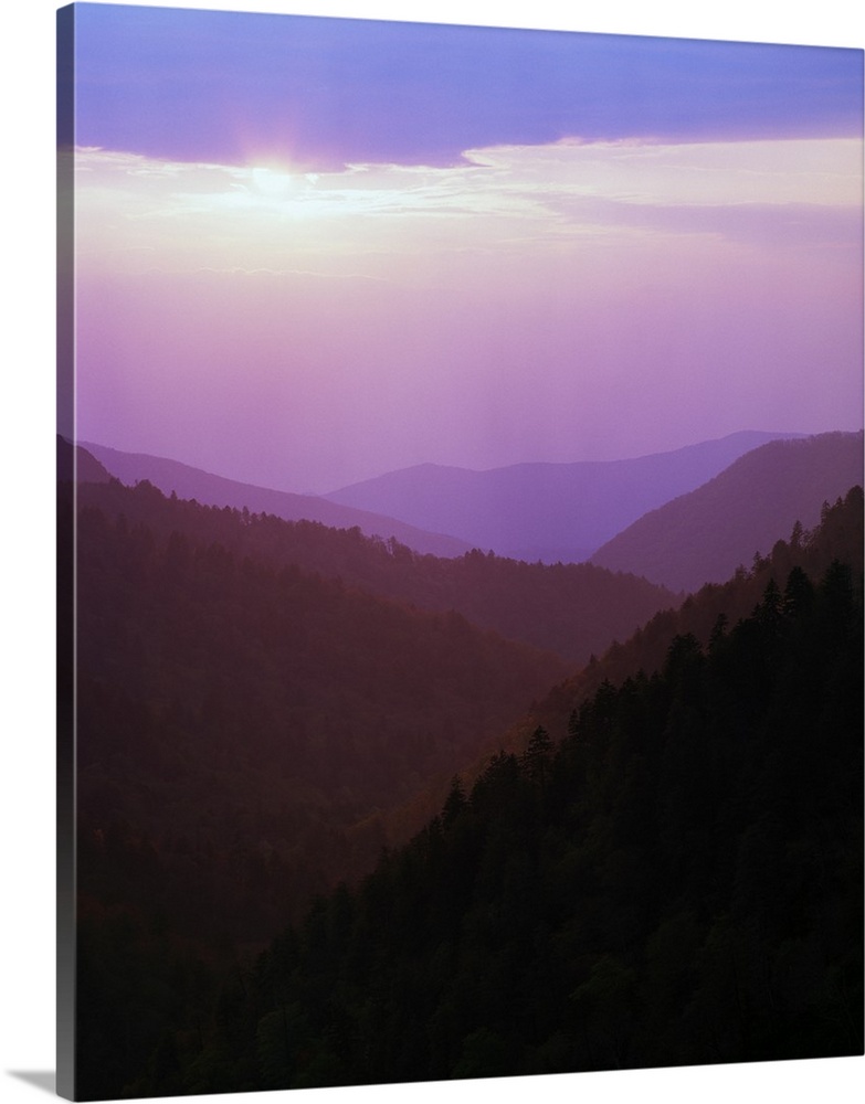 Photograph of foggy mountain range under a cloudy sky with sun peeking through.