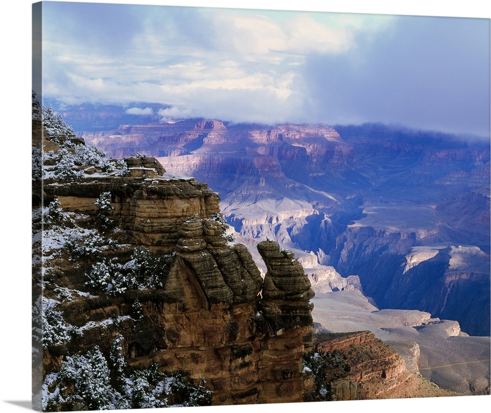 Thick clouds hover over the Grand Canyon that is photographed from a distance behind a snowy cliff.