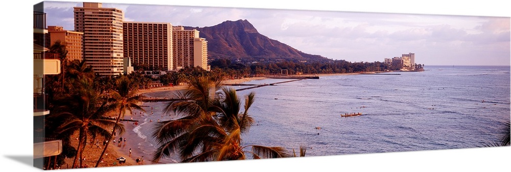 A Hawaiian coast is photographed in wide angle view with a hotel sitting on the beach to the left and a large mountain sho...