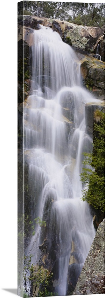This tall waterfall is pictured in an elongated view as it flows over large rocks.