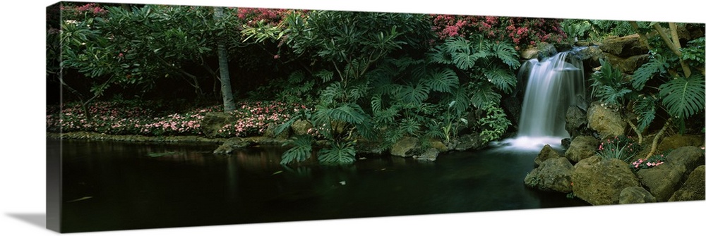 A panoramic photograph of a tropical garden, small cascade tumbles into a circular pool lined with palm fronts and small t...