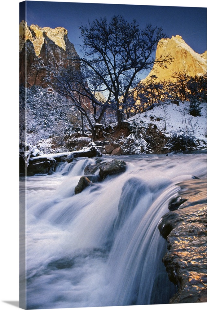 Water rushing over a rocky riverbed in a snowy landscape under bare trees in a valley in Zion National Park, Utah.