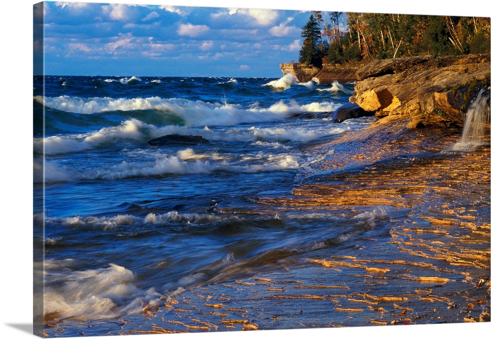 Waves crash onto the rugged lake shore highlighted from the sunset with layered clouds in the distance in this panoramic i...
