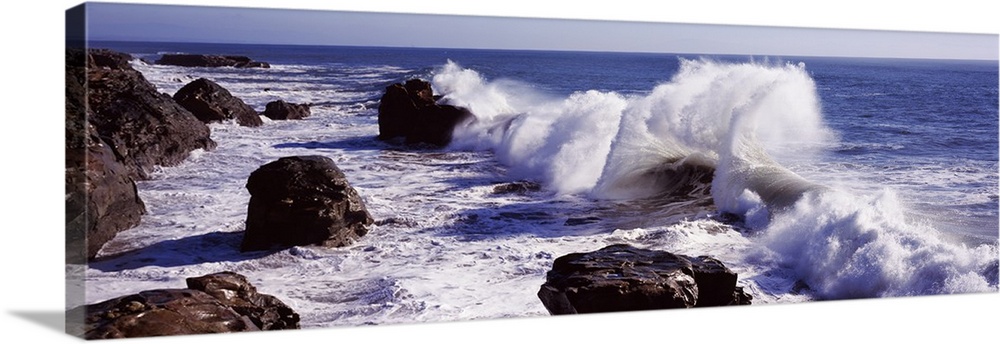 Panoramic photograph of rock cliff line with large boulders and crashing waves under a clear sky.