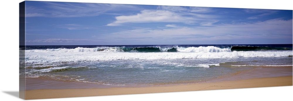 Several clouds in the sky over crashing foamy waves on a sandy beach on the West Coast in a panoramic view.