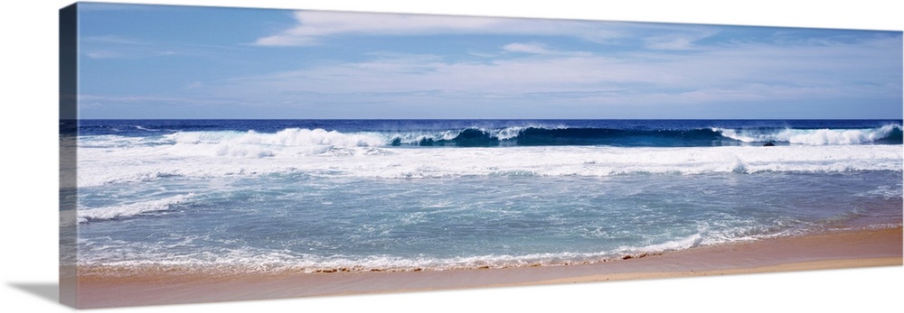 Panoramic photograph displays the waves of an ocean aggressively crashing into a sandy beach on a sunny day.