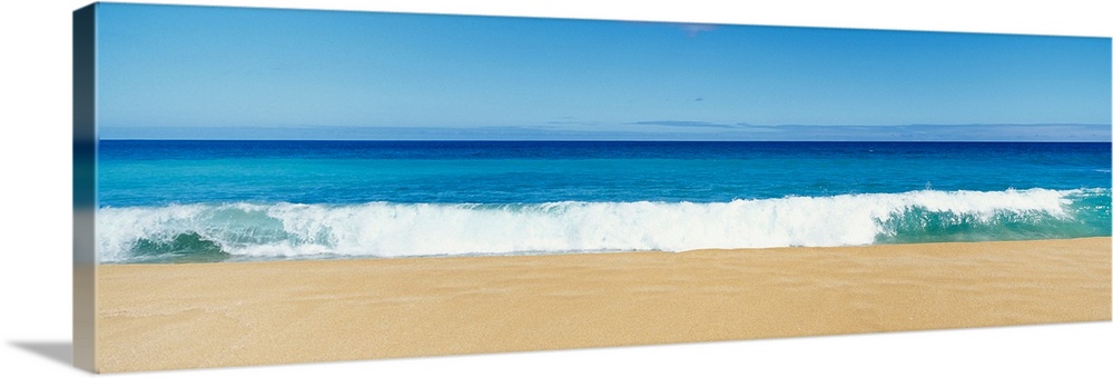This panoramic photograph captures a swell breaking on the sandy shore of a tropical beach on a clear and sunny day.