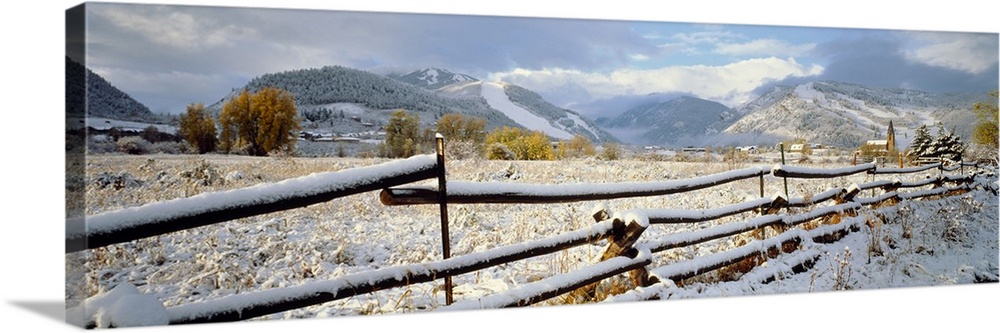 Panoramic photo of a snow covered landscape with a thin wooden fence running through it in the foreground and rolling moun...