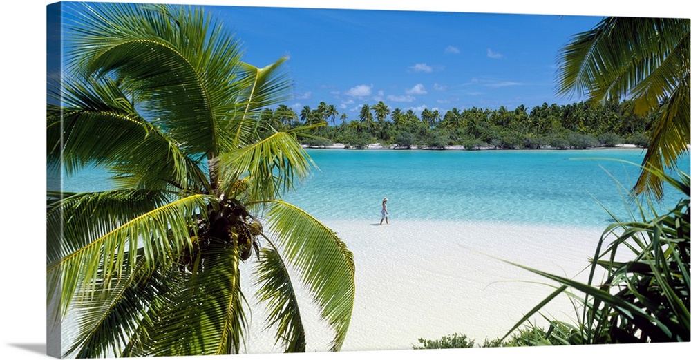 Photograph of a lady walking alone on a white sand beach surrounded by crystal clear water and palm trees.