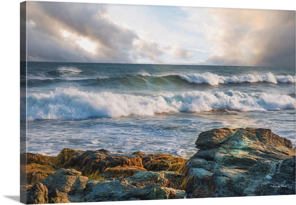Rolling waves in the ocean, seen from the rocky shore, with pastel clouds overhead.