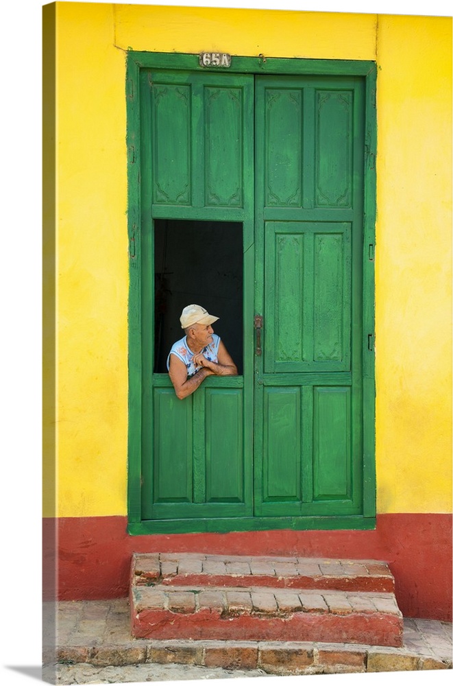 Photograph of a man leaning through a window connected to a large green door on a yellow and red wall in Cuba.