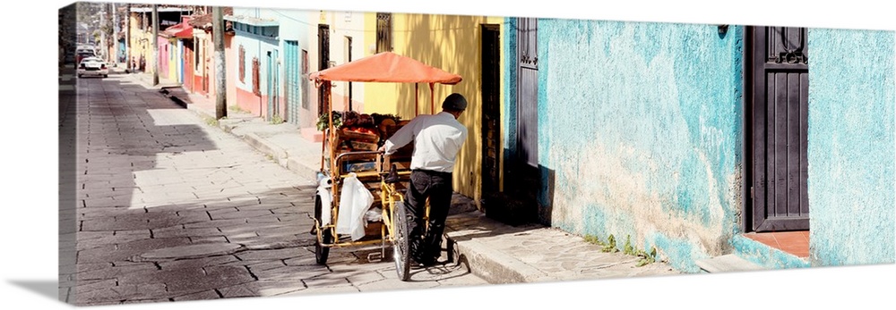 Panoramic photograph of a street vendor pushing his cart full of fresh fruit down a colorful street in Mexico. From the Vi...