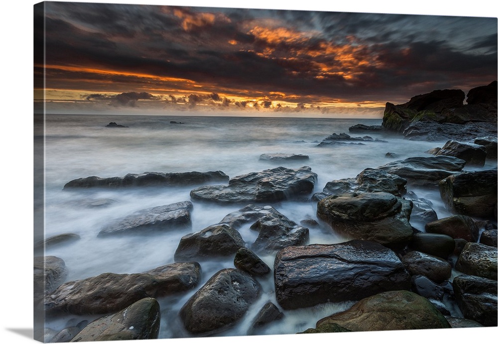 Dramatic sky filled with dark clouds illuminated by the setting sun, with a rocky coastline in the foreground.