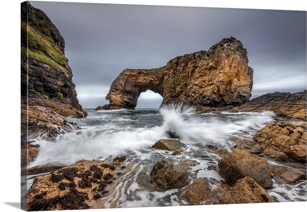 Oceanwater splashing onto the rocky shores in Ireland, with a natural bridge formation in the distance.