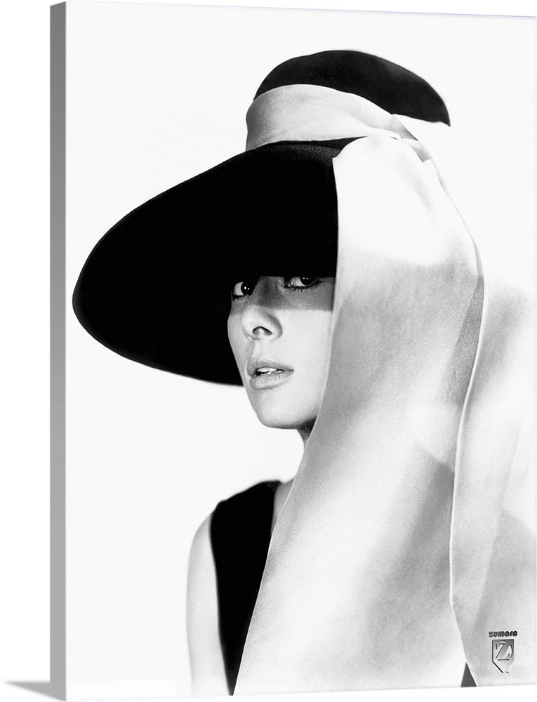 Black and white photo of Audrey Hepburn in a black dress and large floppy black hat with a white sash tied to the hat stre...
