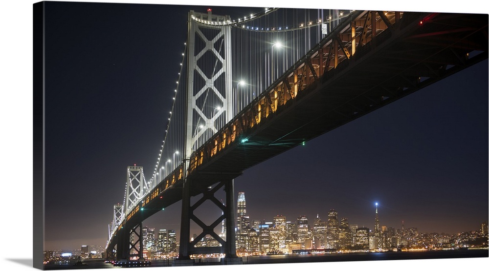 Photograph of the Bay Bridge at night from below with the San Francisco skyline lit up in the background.