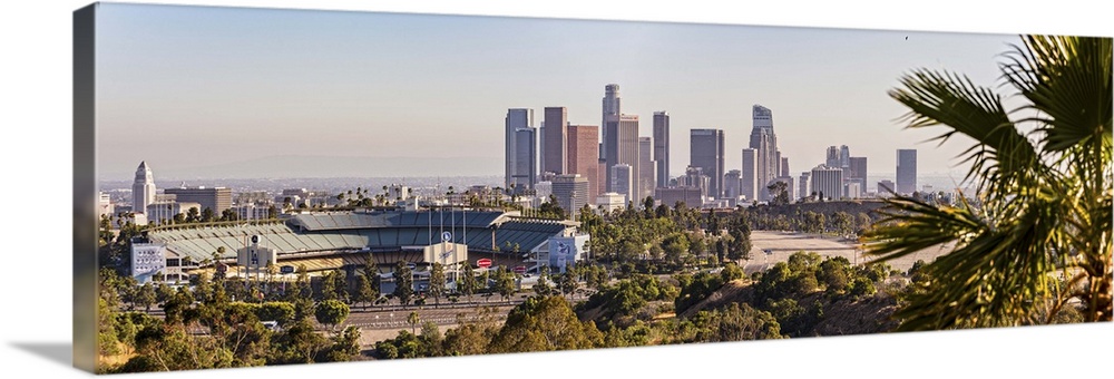 Panoramic photograph of the downtown Los Angeles skyline with Dodger Stadium on the left and a palm tree in the foreground.