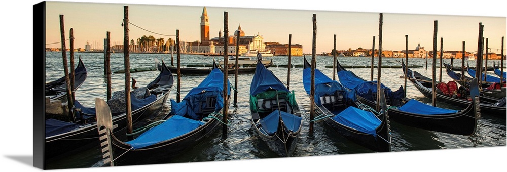 Panoramic photograph of a row of docked gondolas with St. Mark's Square (Piazza San Marco) in the background at golden hour.