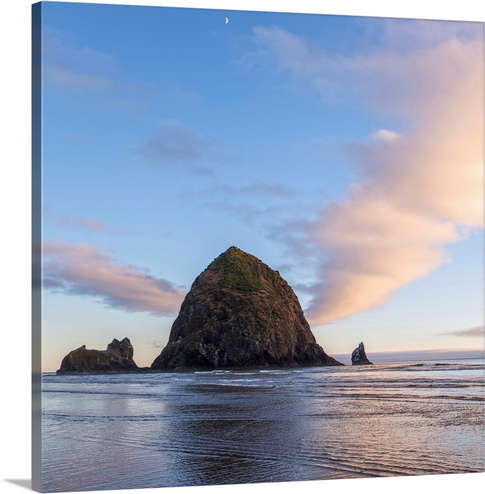 Square photograph of Haystack Rock at sunset with rippling waters in the foreground and the moon above.