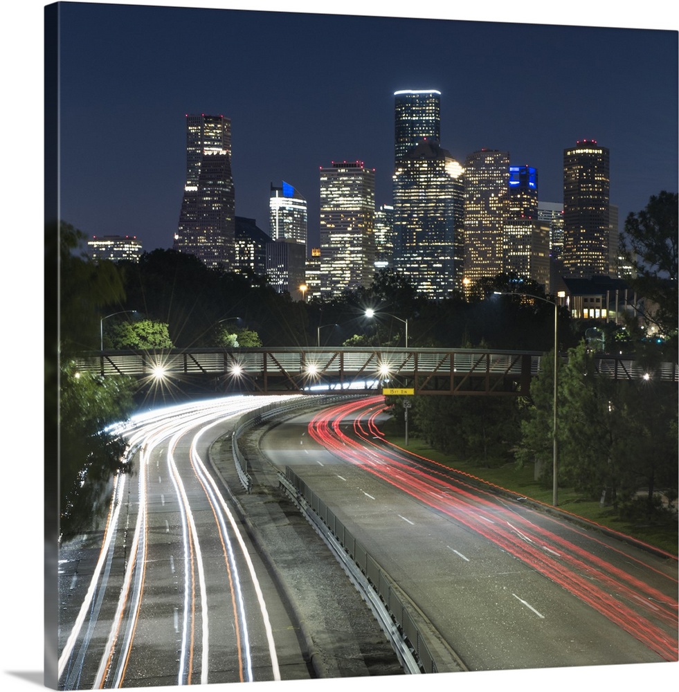 Square photograph of the Houston, TX skyline at night with curving light trails from cars driving by below the Rosemont pe...