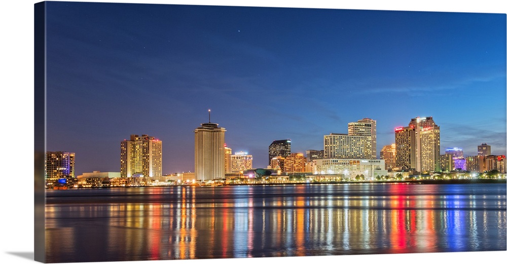 Photograph of the New Orleans skyline lit up at night and reflecting colorful bands onto the Mississippi River.