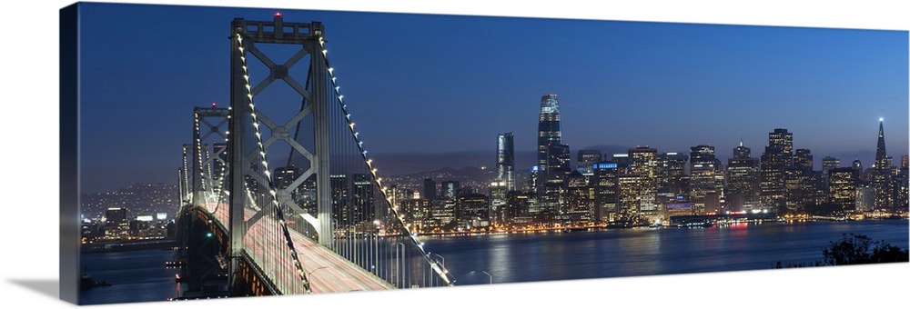 Panoramic photograph of the Bay Bridge and the San Francisco skyline lit up at dusk.