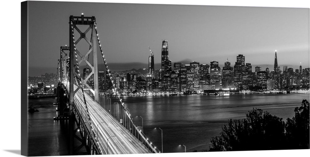 Photograph of the Bay Bridge with a sunset and the San Francisco skyline lit up in the background.