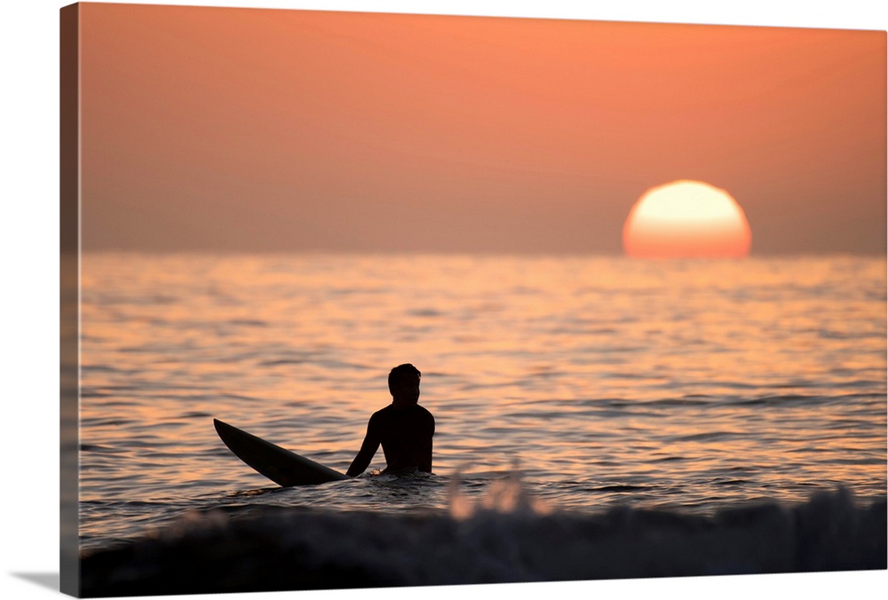 Silhouetted photograph of a man sitting on his surfboard in the Pacific Ocean on the coast of San Diego, California with a...
