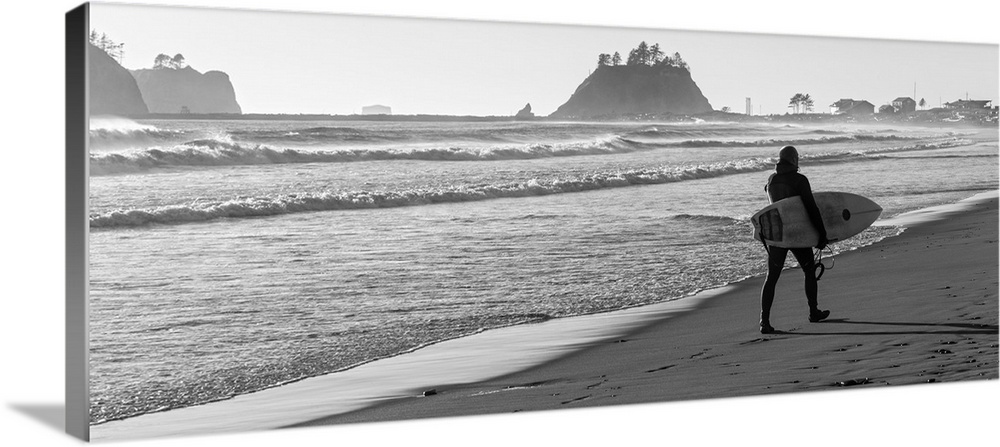Black and white panoramic photograph of a surfer walking along the shore at La Push Beach in Washington.