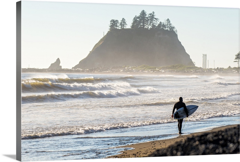 Photograph of a surfer walking on the shore of La Push Beach in Washington, with misty cliffs in the background.