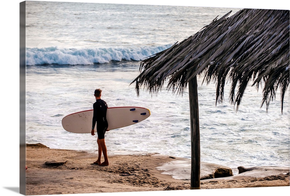 Photograph of a surfer walking along the shore of the pacific ocean in San Diego, California, with a cabana built with nat...