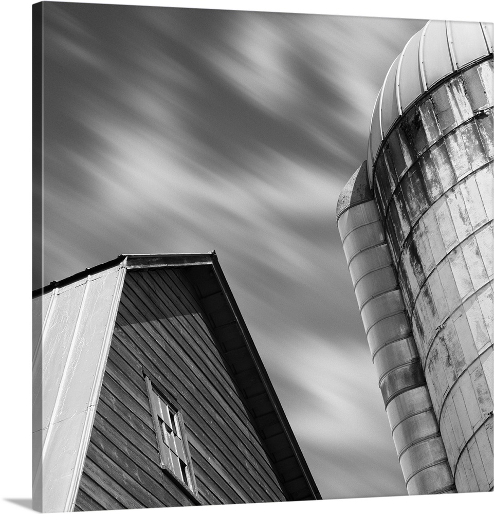 A black and white image of a barn and silo with a blurred clouded sky.