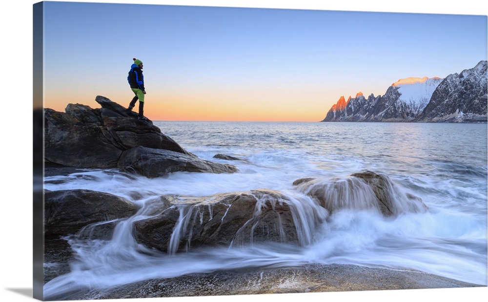 Hiker admires the waves of the icy sea crashing on the rocky cliffs at dawn, Tungeneset, Senja, Troms county, Arctic, Norw...