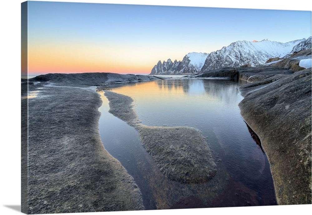 Orange sky at sunset reflected on snowy peaks and the frozen sea surrounded by rocks Tungeneset, Senja, Troms County, Arct...